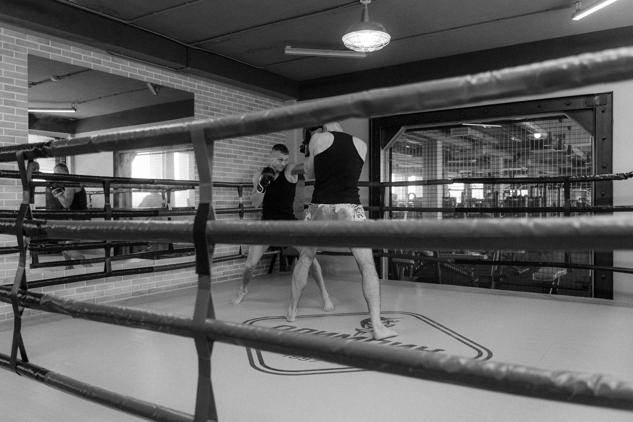 Black and white image of two men sparring in a boxing ring inside a gym.